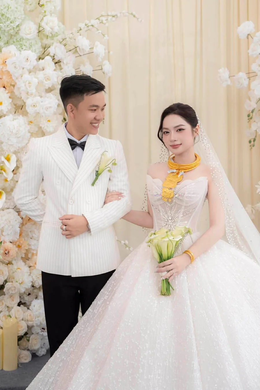 Chú rể vest trắng và cô dâu váy cưới bên phông hoa trắng