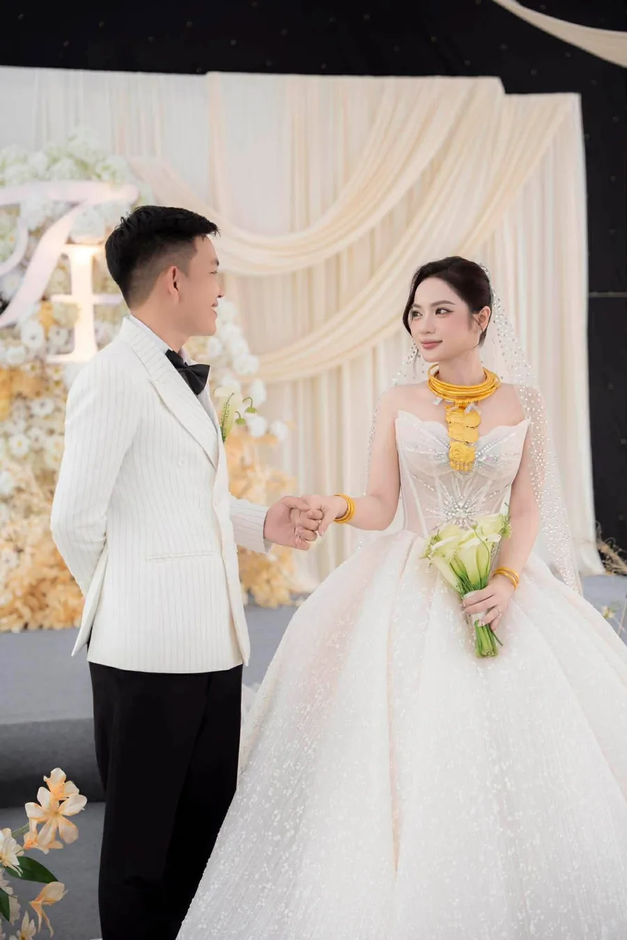 Cô dâu chú rể nắm tay nhau trước phông màn kem